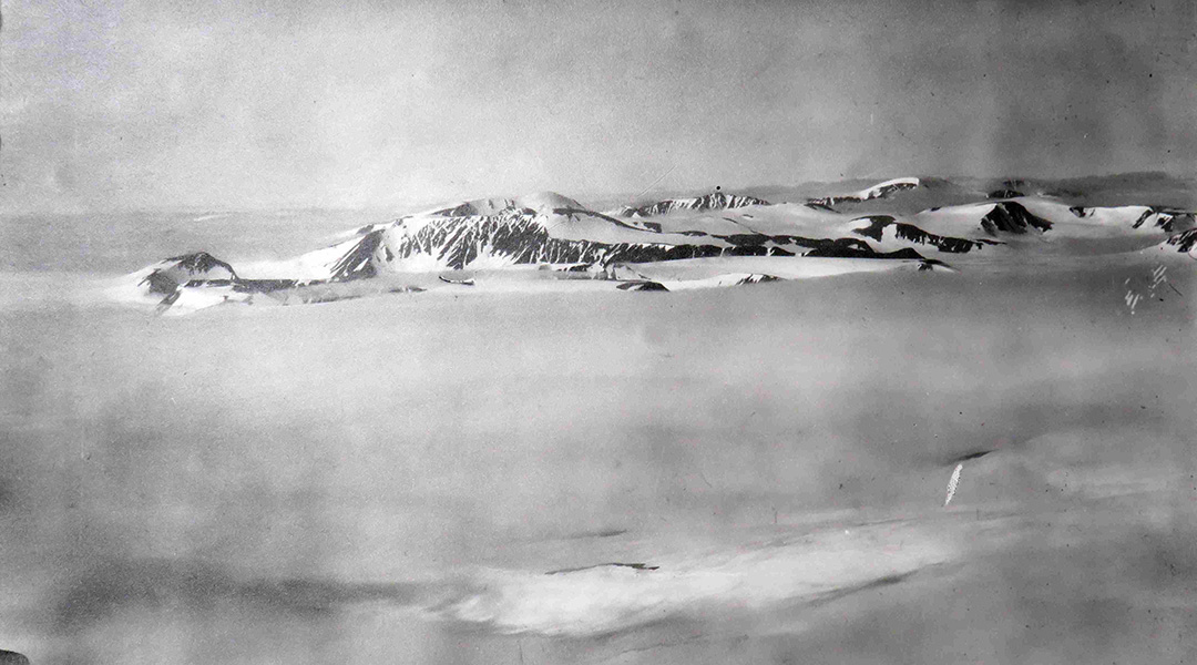 Schwarzweiß Schrägluftbild einer weiten Schneelandschaft mit verschneitem Bergkamm.